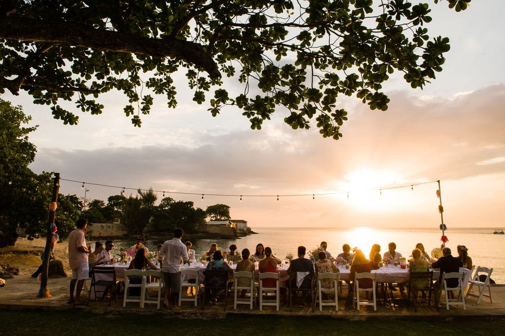 Guests at a long dinner table by the water at sunset