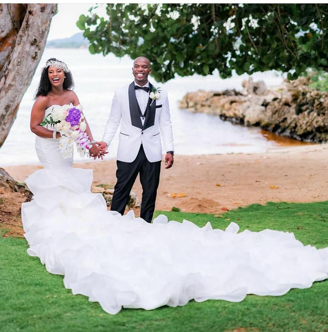 A smiling bride and groom hand in hand on the lawn by the beach, her long ruffled train spread across the grass