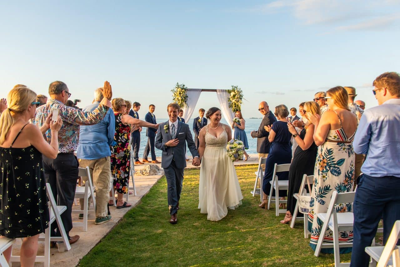 The newlyweds walking back down the aisle past cheering guests, with the flower arch and the sea behind them