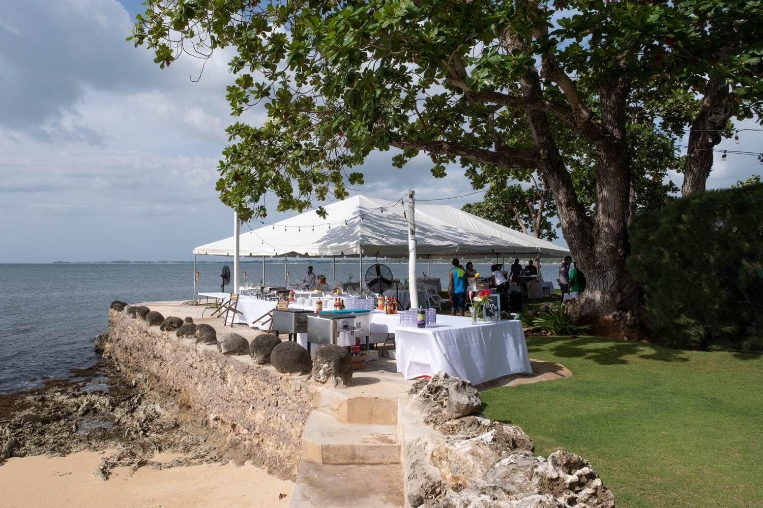 A white tent set up on the lawn beside the seawall, under a large tree