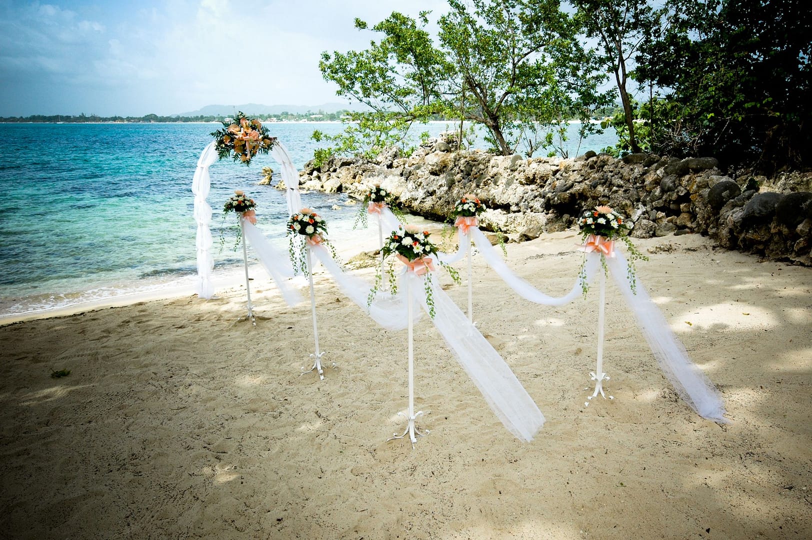 A white wedding arch dressed with flowers and fabric on the sand, facing the sea