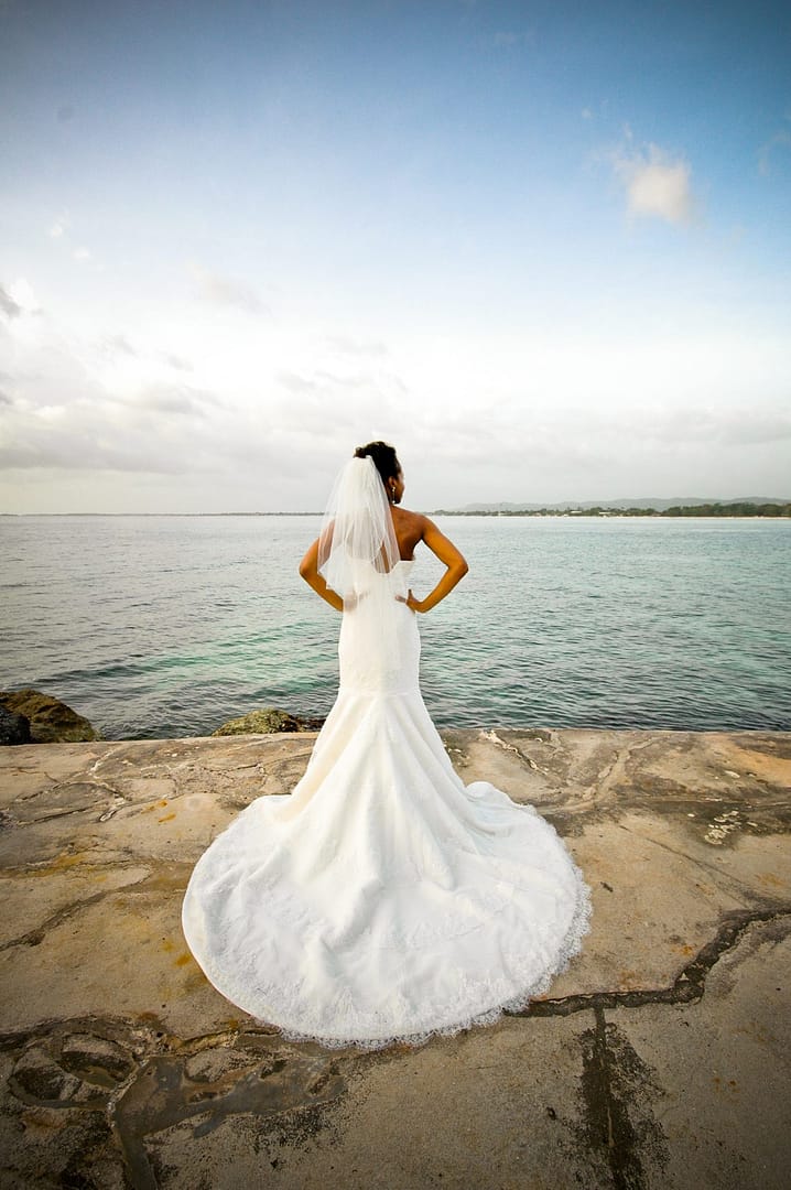 A bride in a long-trained gown standing on the stone seawall, looking out over the bay